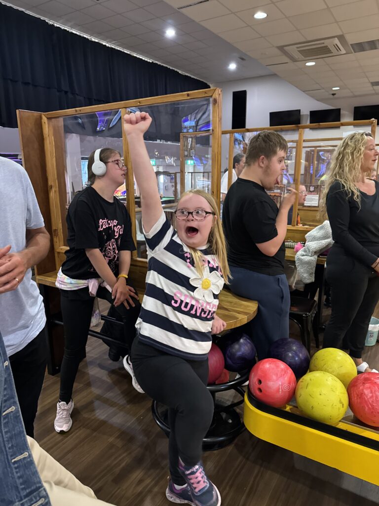 A young girl with Down syndrome strikes a triumphant pose at a bowling alley, arm raised in celebration. She’s wearing a striped T-shirt with a flower and the word “Sundown” on it. Around her, other young people and adults are engaged in the activity, with colourful bowling balls visible nearby. The setting is lively and inclusive, full of energy and joy.