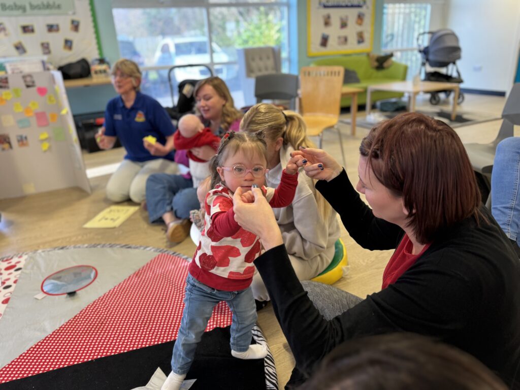 A young girl with Down's Syndrome wearing heart-print top and glasses is supported while standing during a Sundowns baby group session. Other parents and children sit around on the floor in a bright and welcoming room, engaging in activities together.