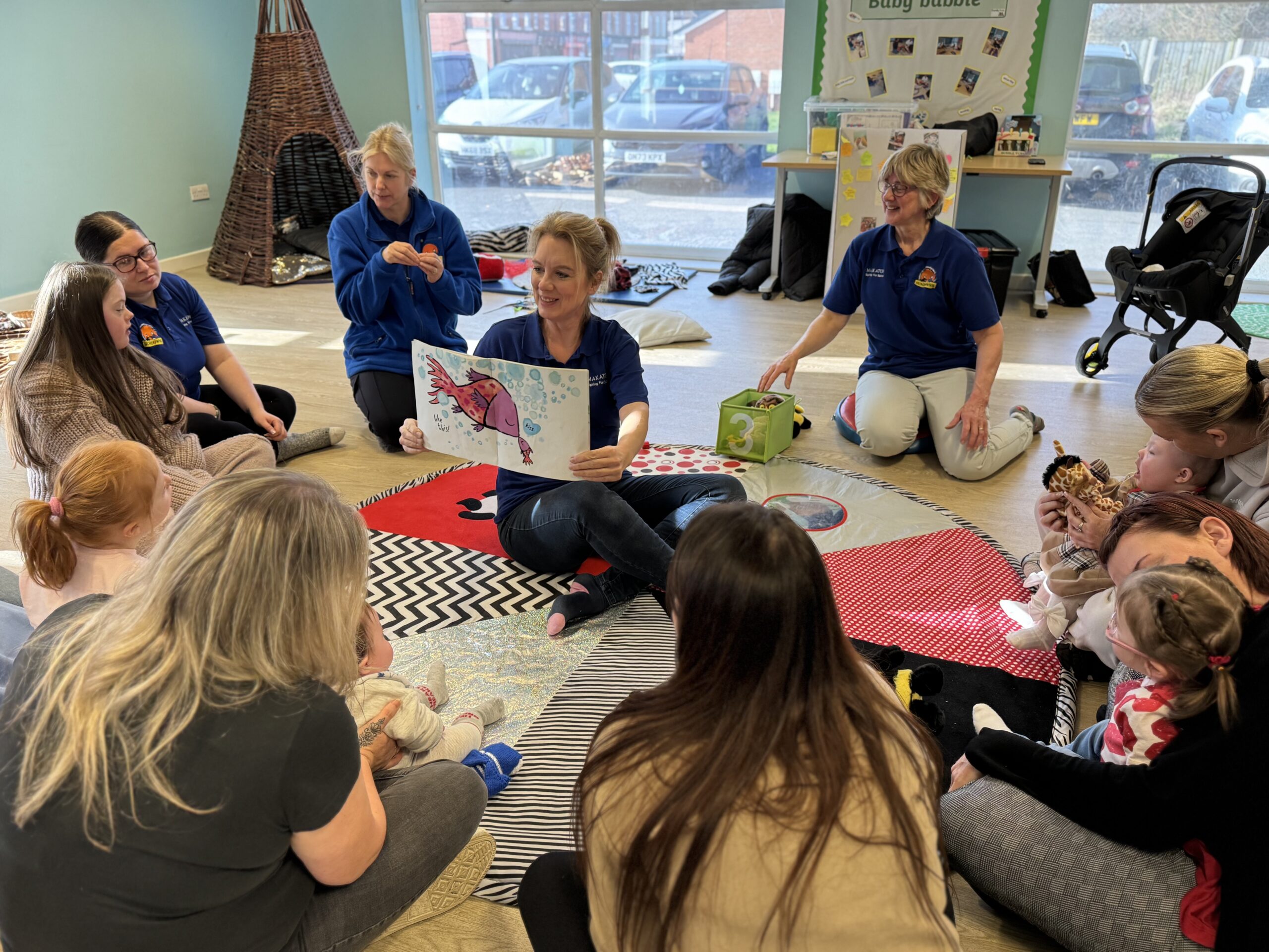 A group of parents and children, including a girl with Down syndrome, sit in a circle on a colourful sensory mat as a woman reads a storybook. Three staff members in matching blue polo shirts lead the interactive session.
