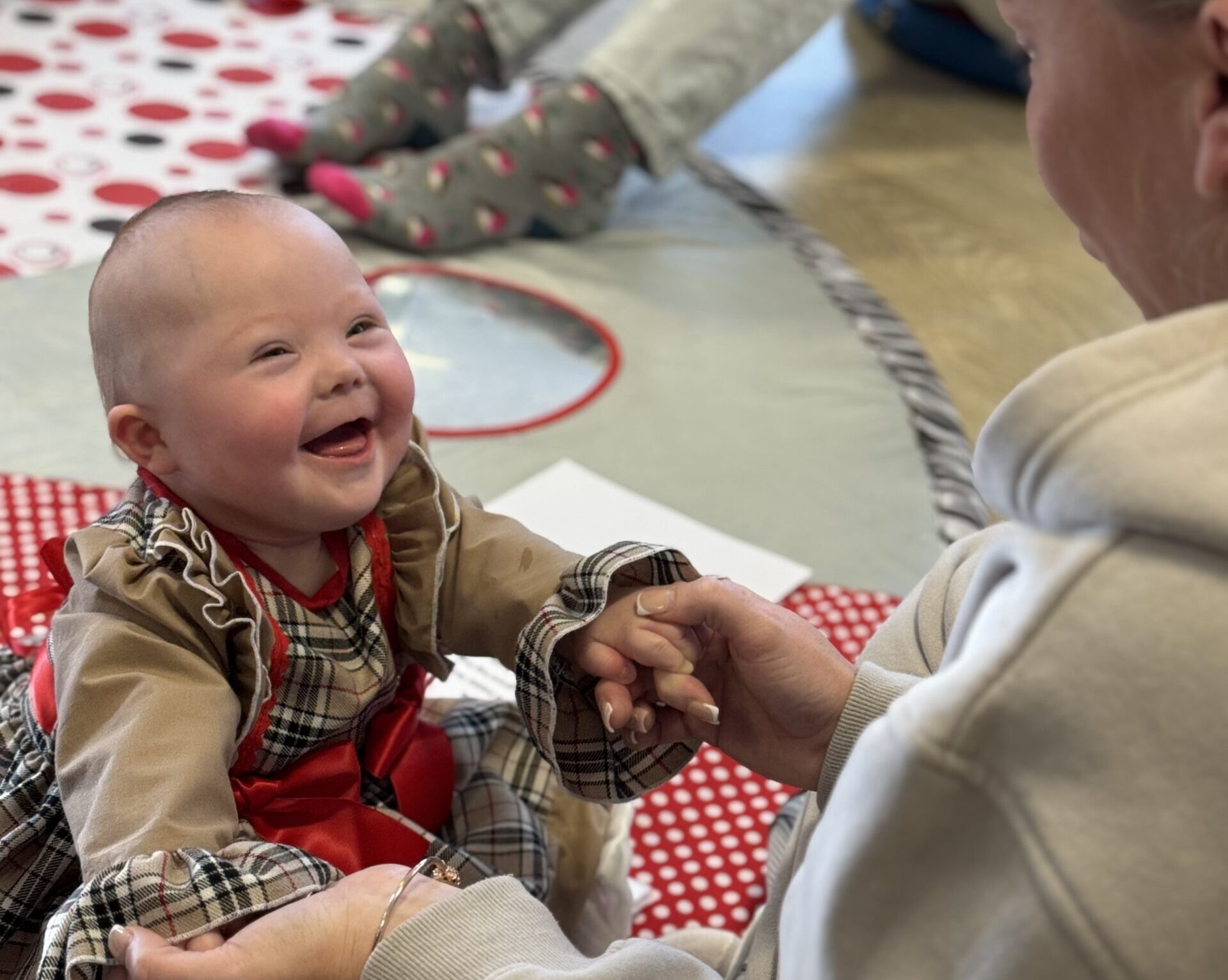 A smiling baby with Down syndrome wearing a tartan dress holds hands with an adult during a Sundowns baby group session on a soft play mat.