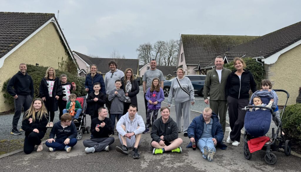 A large group of Sundowns families smiling together outdoors on a residential strip to Ribby Hall. The group includes children, young people, and adults, some of whom have Down syndrome. Several people are seated on the ground, while others stand or use pushchairs. Everyone appears relaxed and happy, with Ribby Hall houses in the background.
