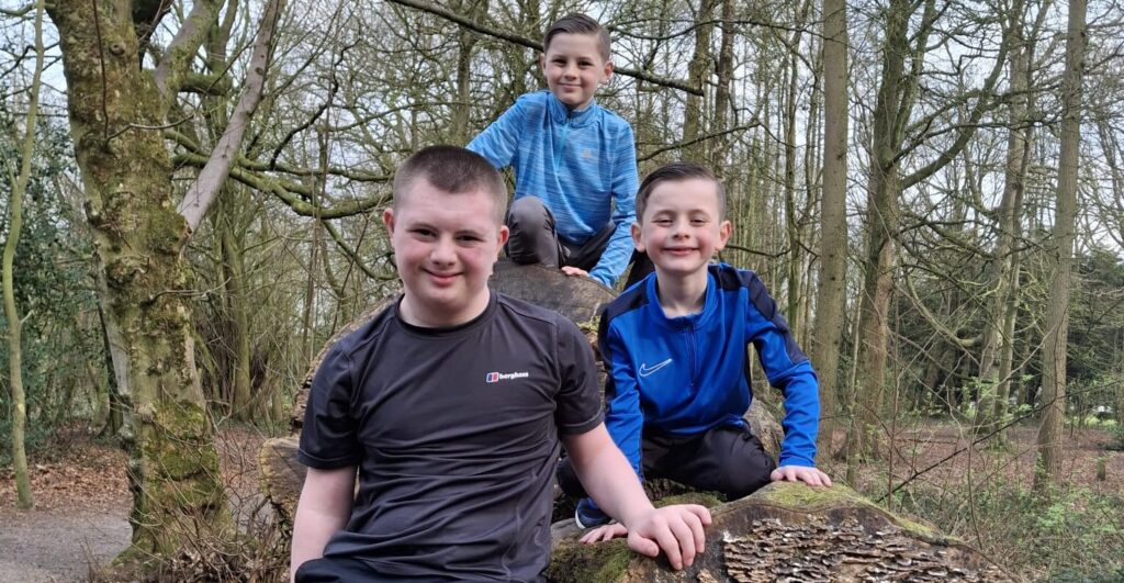 Three brothers, including one with Down syndrome, smiling and climbing on a fallen tree in a woodland at Ribby Hall.