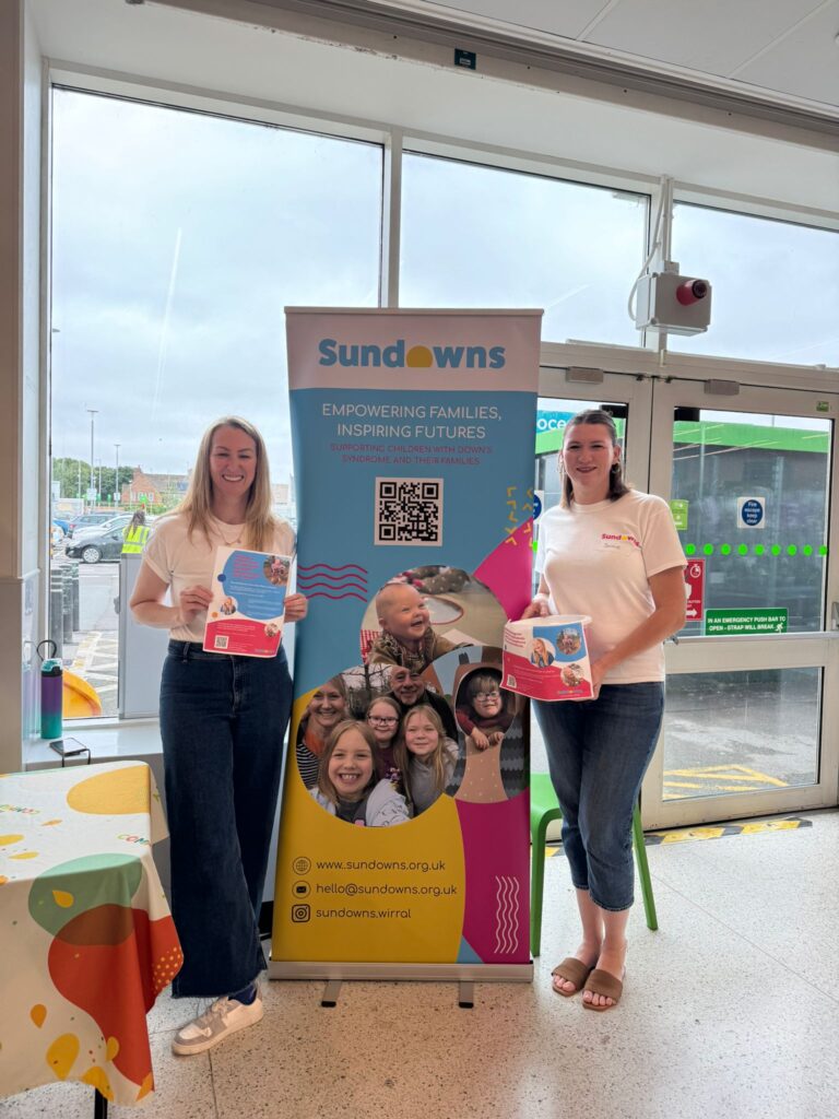 Two women stand smiling on either side of a vibrant Sundowns promotional banner inside a supermarket entrance. The banner features the Sundowns logo and tagline "Empowering Families, Inspiring Futures," and highlights the charity’s work supporting children with Down syndrome and their families. The banner displays colourful shapes, joyful photos of children and families, a QR code, and Sundowns' contact details. Both women are holding leaflets related to Sundowns.