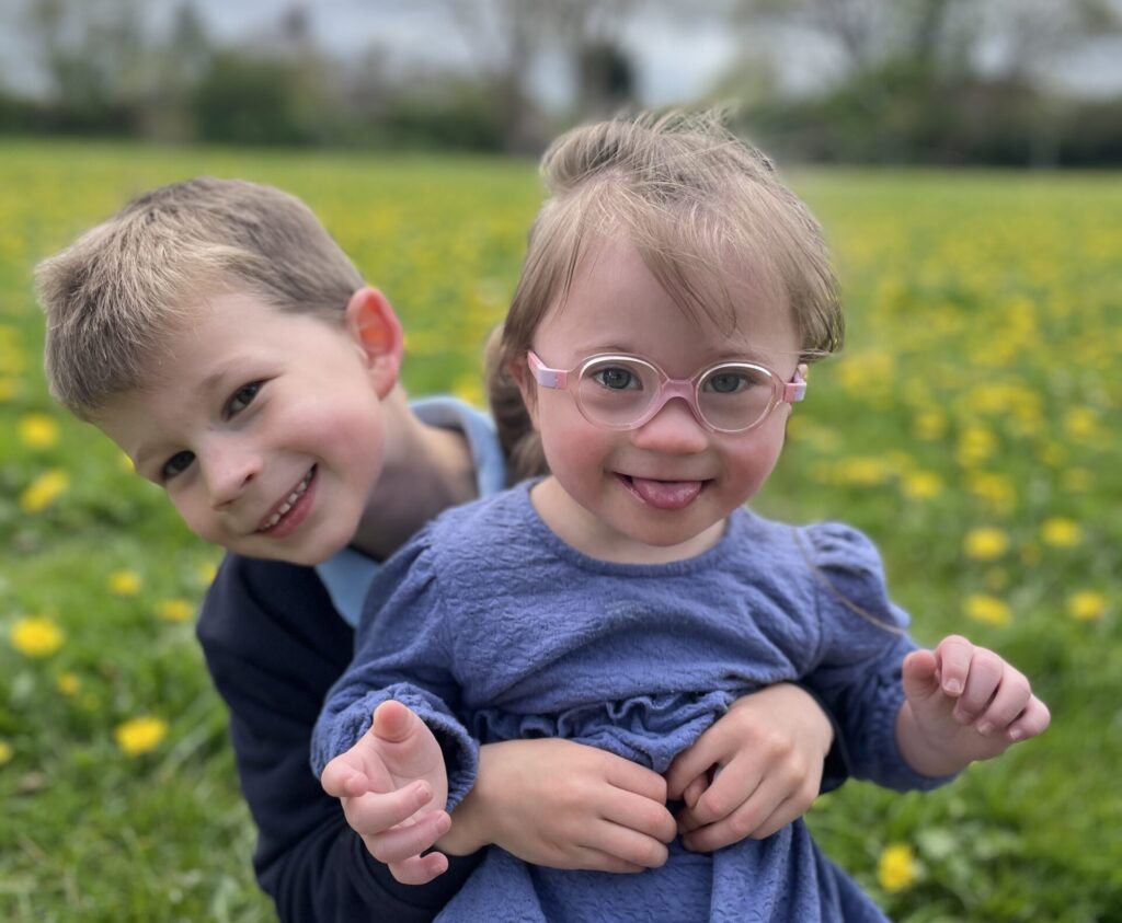 A young girl with Down syndrome wearing pink glasses is lovingly held by her smiling older brother in a field full of yellow flowers.